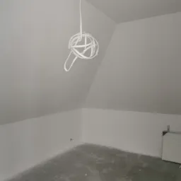 Poddasze po remoncie, świeżo pomalowane na biało, z widocznymi śladami szpachli na podłodze i wiszącą instalacją elektryczną w kształcie kuli.