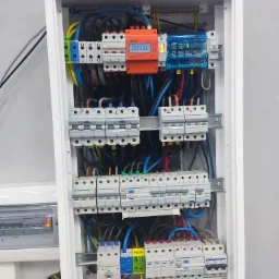 Otwarta skrzynka elektryczna z bezpiecznikami, przewodami w różnych kolorach, zabezpieczeniami przeciwprzepięciowymi i licznikiem energii, zamontowana na ścianie.