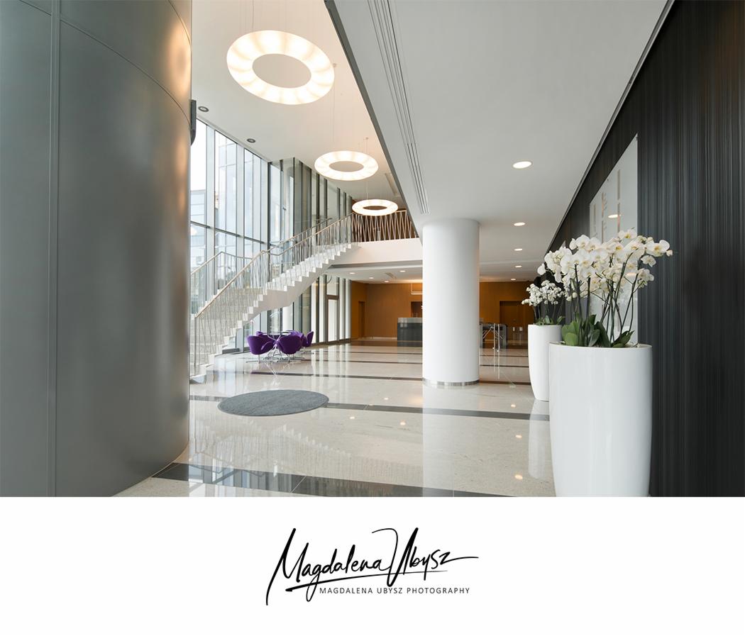 Jasne, przestronne wnętrze biurowca z marmurową podłogą, białymi kolumnami, schodami z metalową balustradą i fioletowymi krzesłami w tle, oświetlone okrągłymi lampami sufitowymi i ozdobione białymi...