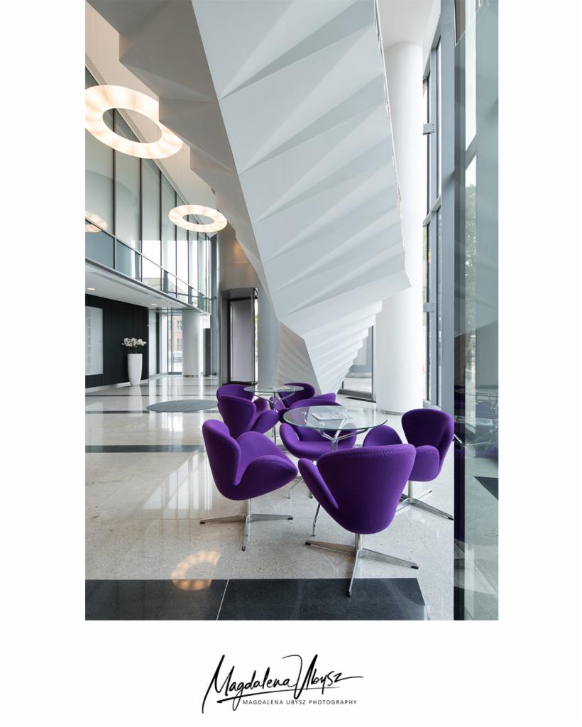 Nowoczesne wnętrze biurowe z fioletowymi krzesłami tulipanowymi wokół szklanego stolika, podwieszany sufit o geometrycznej strukturze, duże okna z widokiem na zewnątrz, marmurowa podłoga.