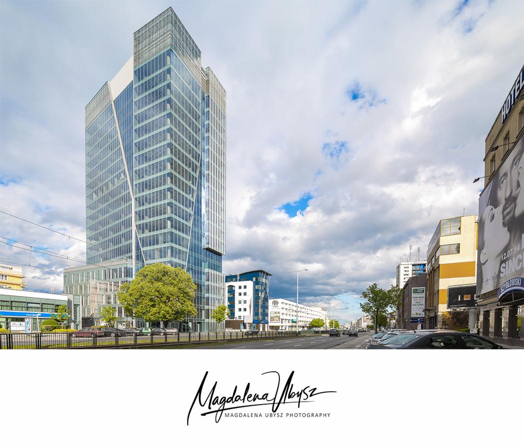 Szklany biurowiec z geometrycznymi podziałami fasady, widoczny z poziomu ulicy z drzewem w pierwszym planie i pochmurnym niebem w tle.