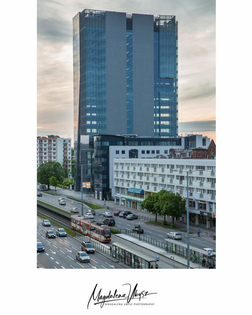 Wysoki, nowoczesny biurowiec z szklaną fasadą górujący nad miejskim krajobrazem, z ulicą, tramwajem i przystankiem w kadrze.