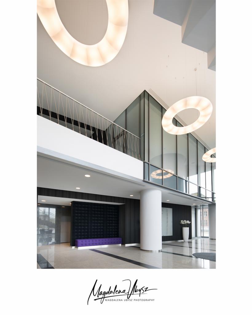 Przestronne, nowoczesne wnętrze biurowca z marmurową podłogą, okrągłymi lampami sufitowymi, antresolą z metalową balustradą i fioletową sofą pod ciemną ścianą z pikowaniem.