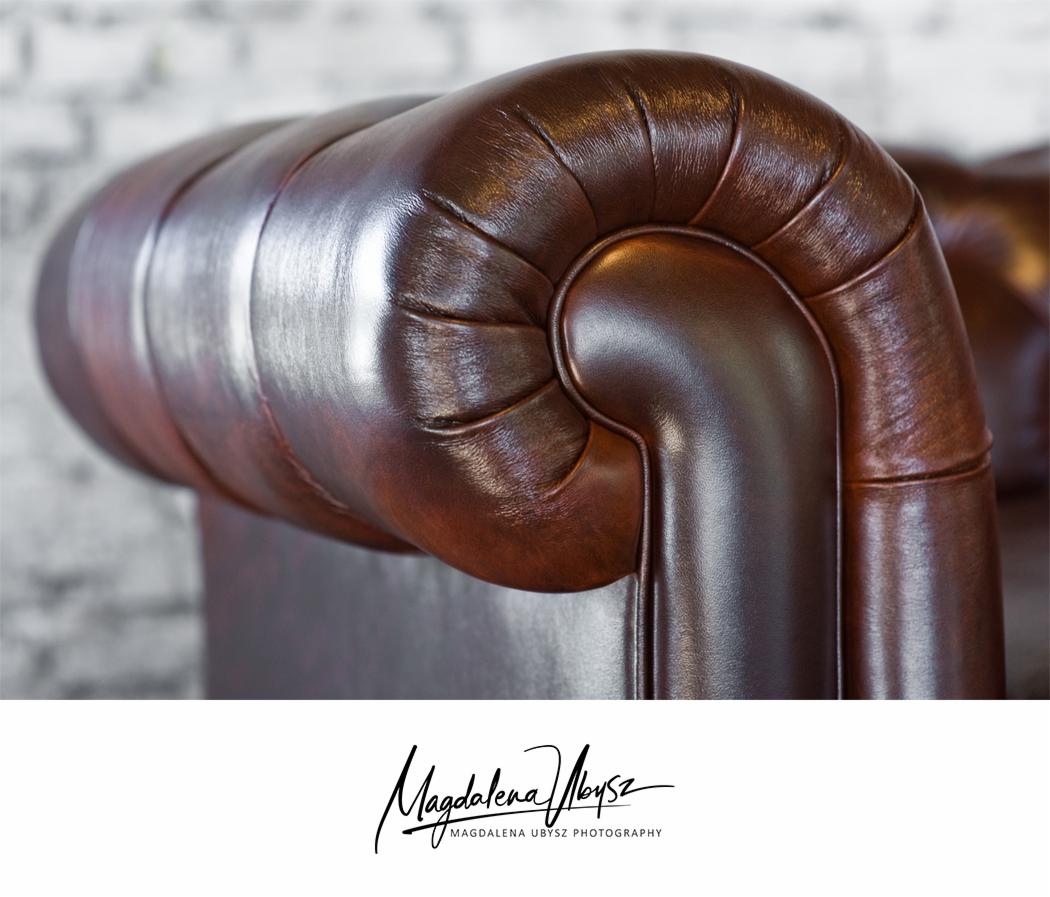 Detal skórzanego, brązowego fotela typu chesterfield z charakterystycznymi przeszyciami na tle ściany z białej cegły.