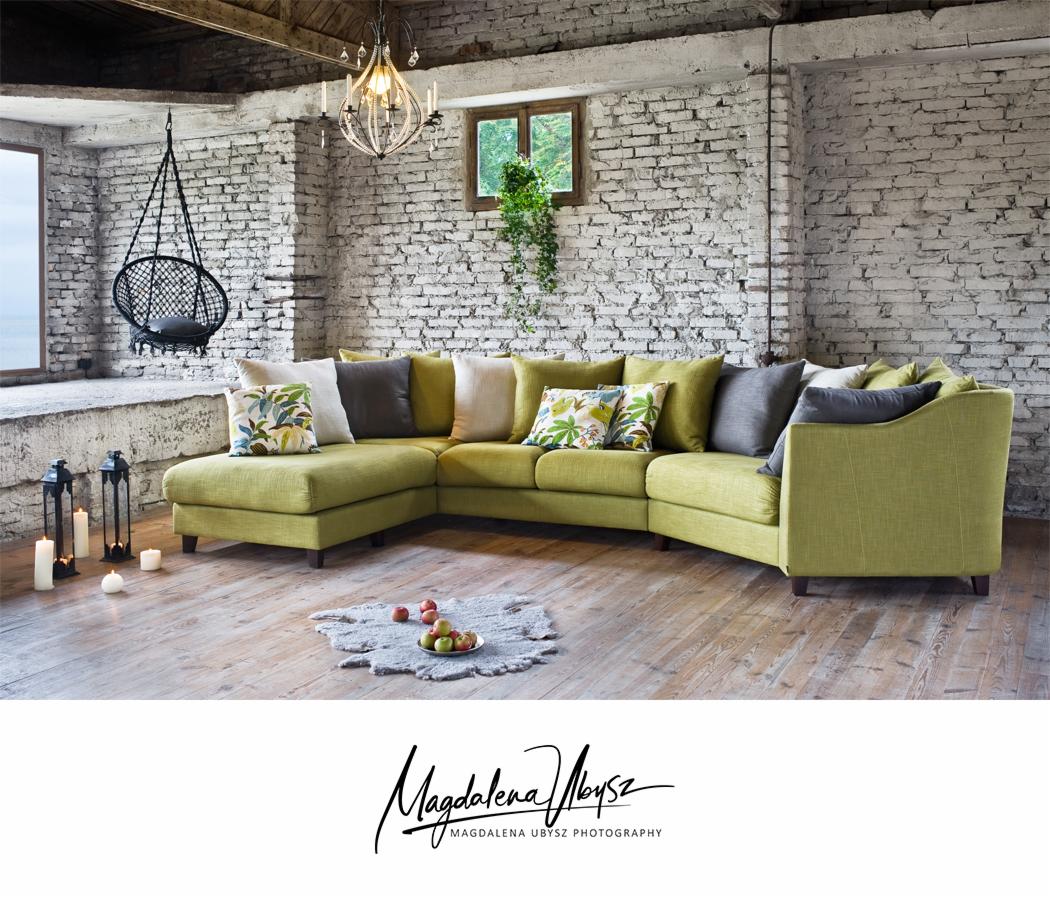 Jasnozielona narożna sofa z poduszkami w loftowym wnętrzu z ceglaną ścianą, wiszącym fotelem i drewnianą podłogą, oświetlona wiszącą lampą, z jabłkami na dywaniku.