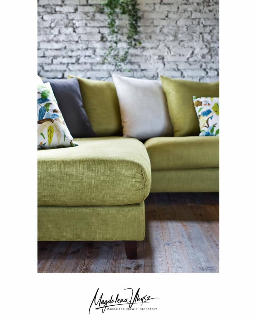 Fragment zielonej, narożnej sofy z poduszkami w odcieniach zieleni, szarości i bieli, ustawionej na drewnianej podłodze na tle białej ściany z cegły i zwisającej rośliny.