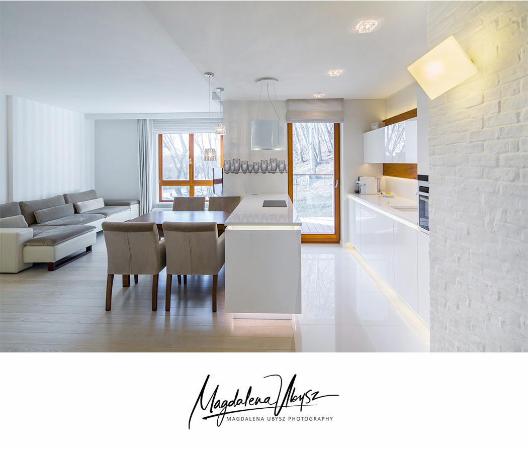 Jasne, nowoczesne wnętrze: salon z sofą i jadalnią płynnie przechodzące w białą kuchnię z połyskiem, ceglana ściana z kwadratową lampą.