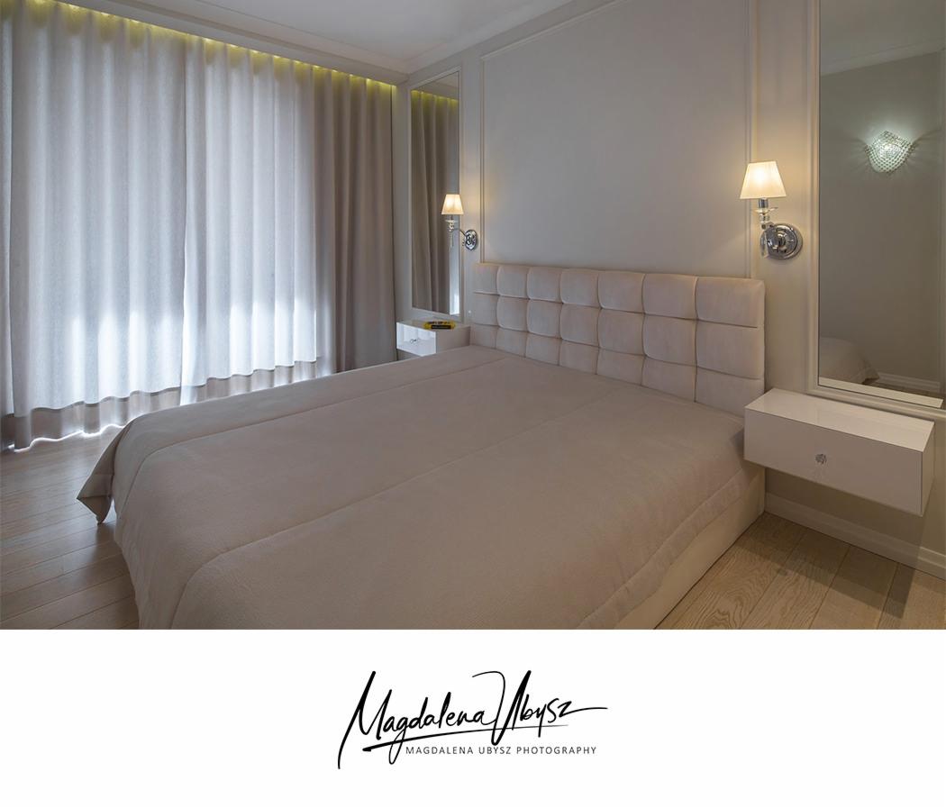 Elegancka sypialnia w jasnych barwach z łóżkiem z pikowanym zagłówkiem, szafkami nocnymi i oświetleniem ściennym, uchwycona w szerokim kadrze.