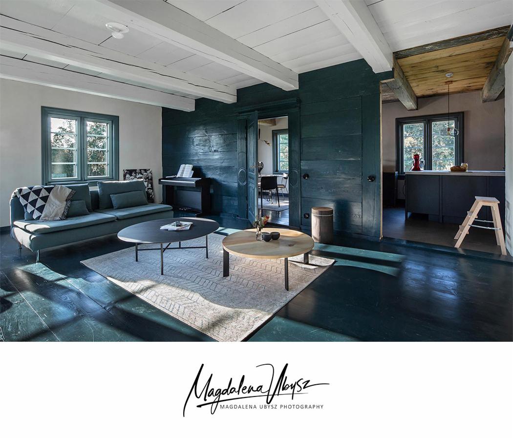 Salon z drewnianą podłogą w odcieniach zieleni, szara sofa z wzorzystymi poduszkami, dwa stoliki kawowe na dywanie, w tle pianino i otwarte drzwi do kuchni.