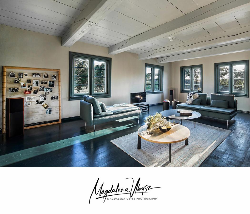 Jasny salon z drewnianą podłogą, sofą, leżanką, stolikami kawowymi i ramą ze zdjęciami, oświetlony wpadającym przez okna światłem słonecznym.