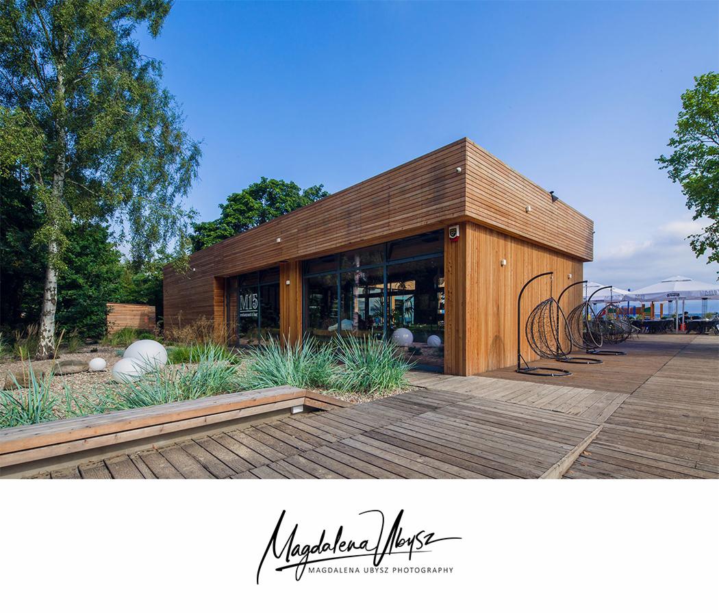 Nowoczesny, drewniany budynek z przeszkloną fasadą, otoczony zielenią i drewnianym tarasem z wiszącymi fotelami. Widoczne elementy krajobrazu: trawy ozdobne, białe kule dekoracyjne, drzewa, niebo.