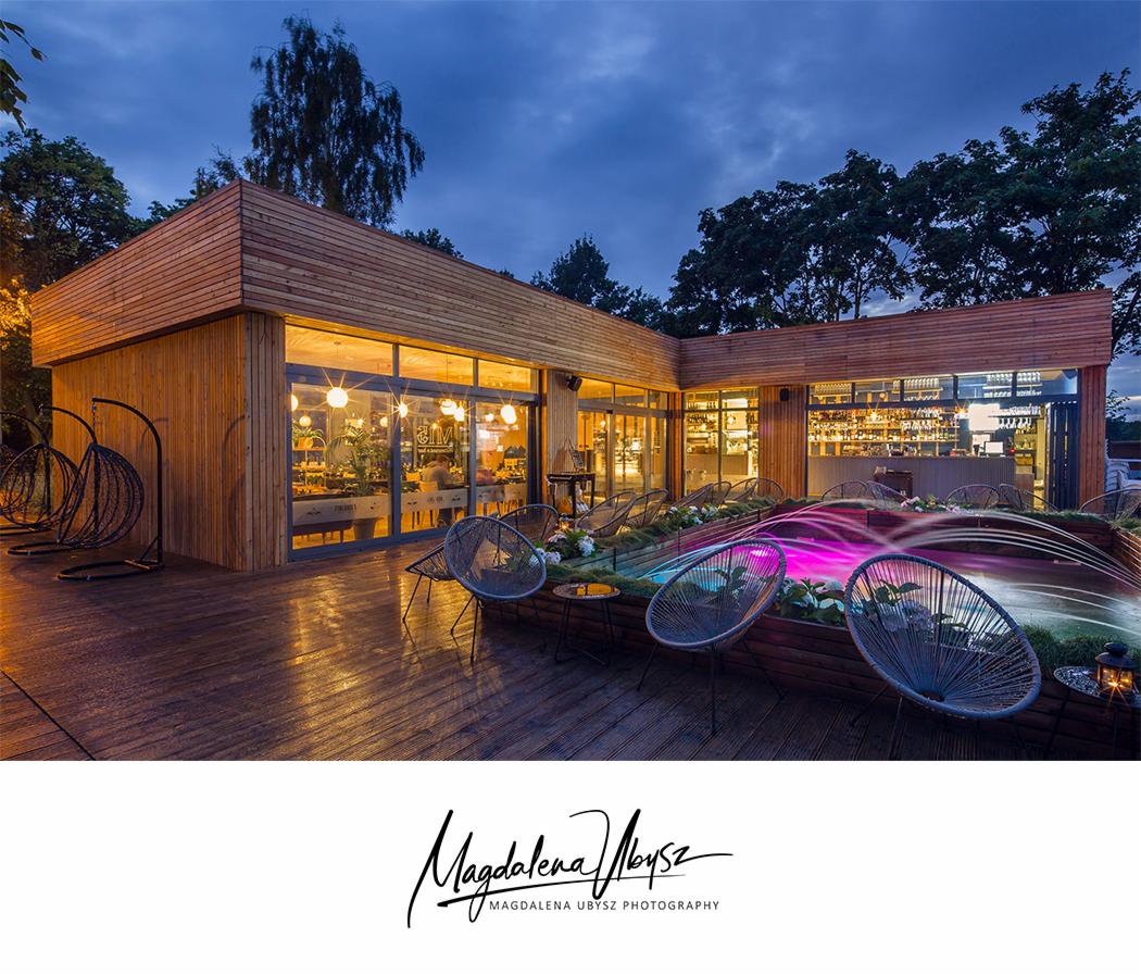Nowoczesny budynek z drewnianą elewacją i dużymi oknami, oświetlony od wewnątrz, z tarasem wyposażonym w plecione krzesła i podświetlaną fontannę, sfotografowany o zmierzchu.