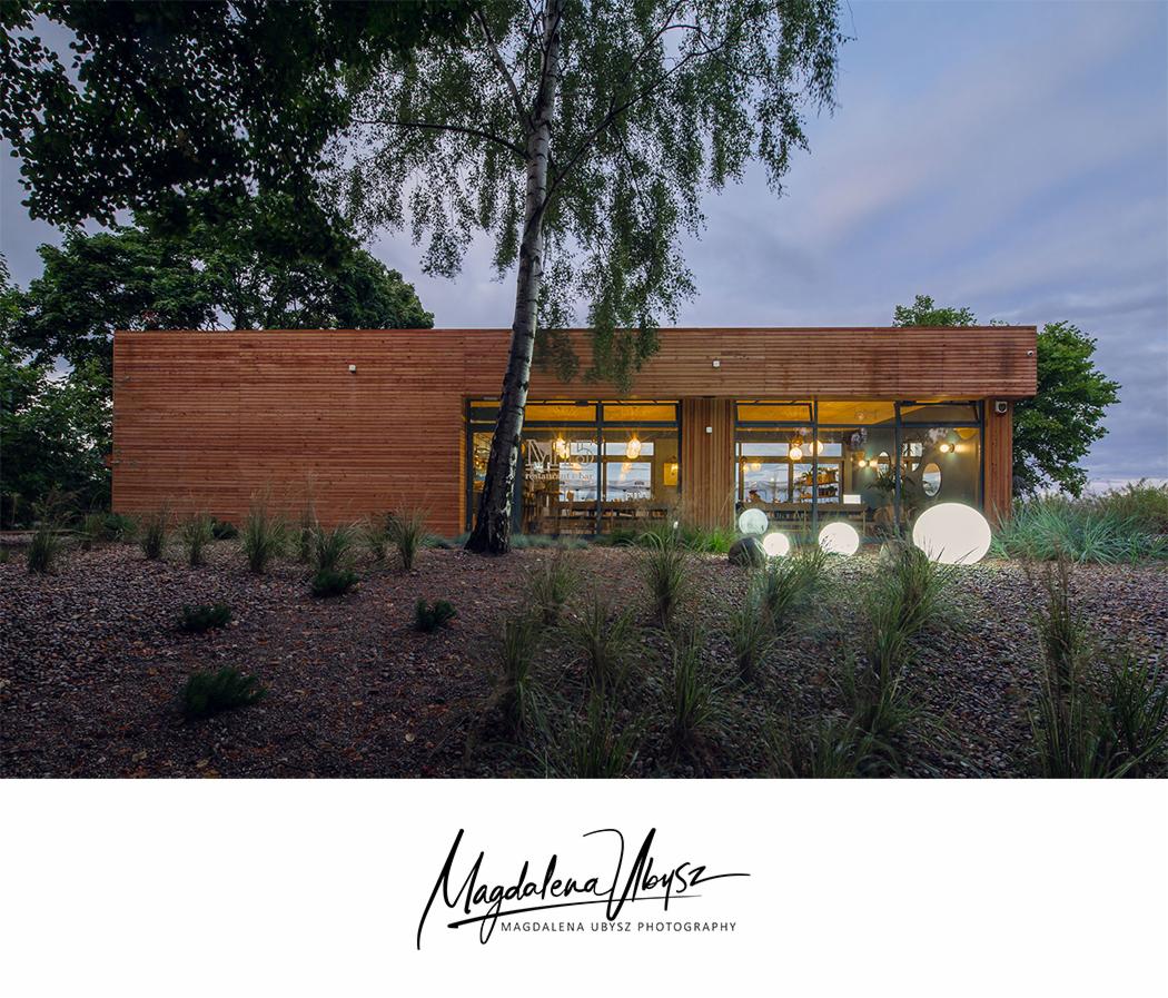 Drewniany budynek restauracji z dużymi oknami i kulistymi lampami na zewnątrz, otoczony zielenią i żwirowym podłożem, sfotografowany o zmierzchu.