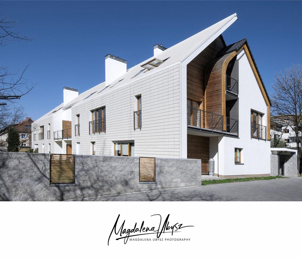 Nowoczesny szeregowiec z białymi panelami elewacyjnymi i drewnianymi akcentami, widok z poziomu ulicy w słoneczny dzień.