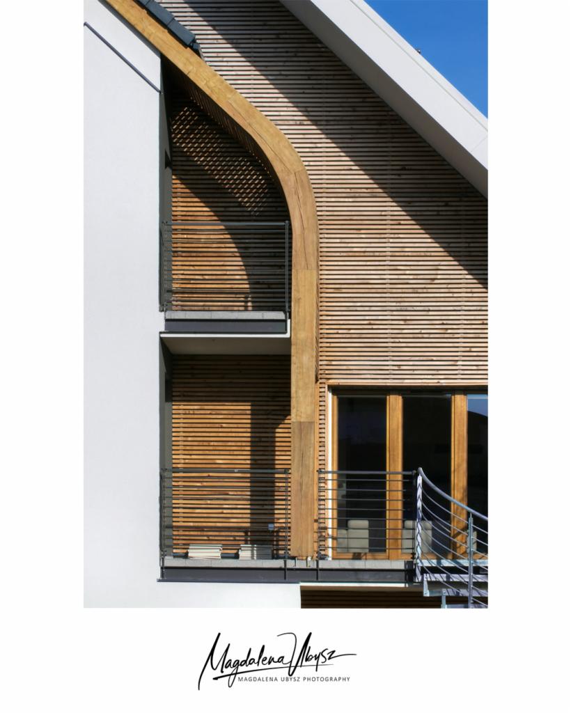 Nowoczesna fasada budynku z drewnianą okładziną, balkonami z metalowymi barierkami i charakterystycznym łukowym elementem konstrukcyjnym z drewna.