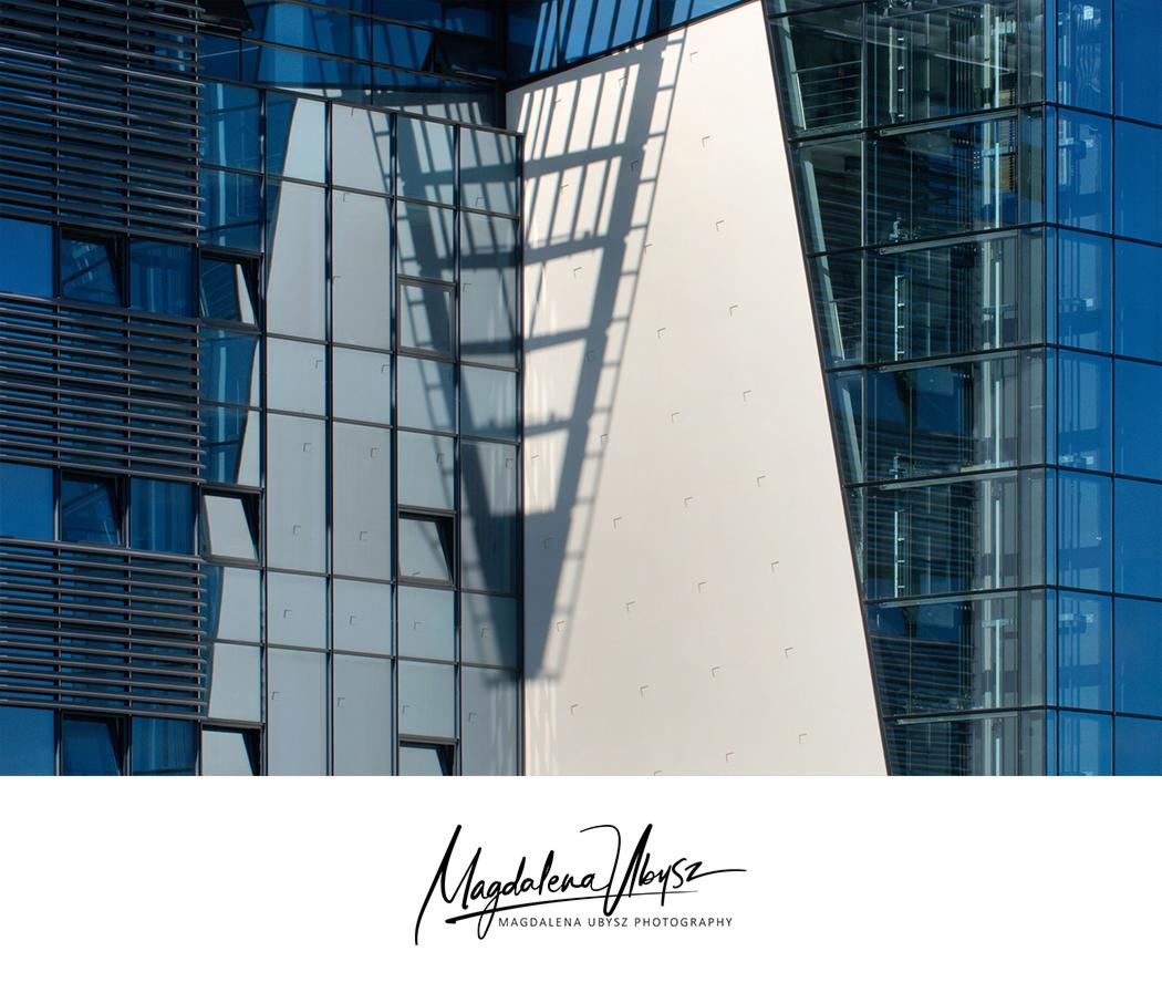 Geometryczne ujęcie fragmentu nowoczesnego biurowca z niebieskiego szkła i białego panelu, na którym widoczne są cienie, z otwartymi oknami i widoczną konstrukcją windy.