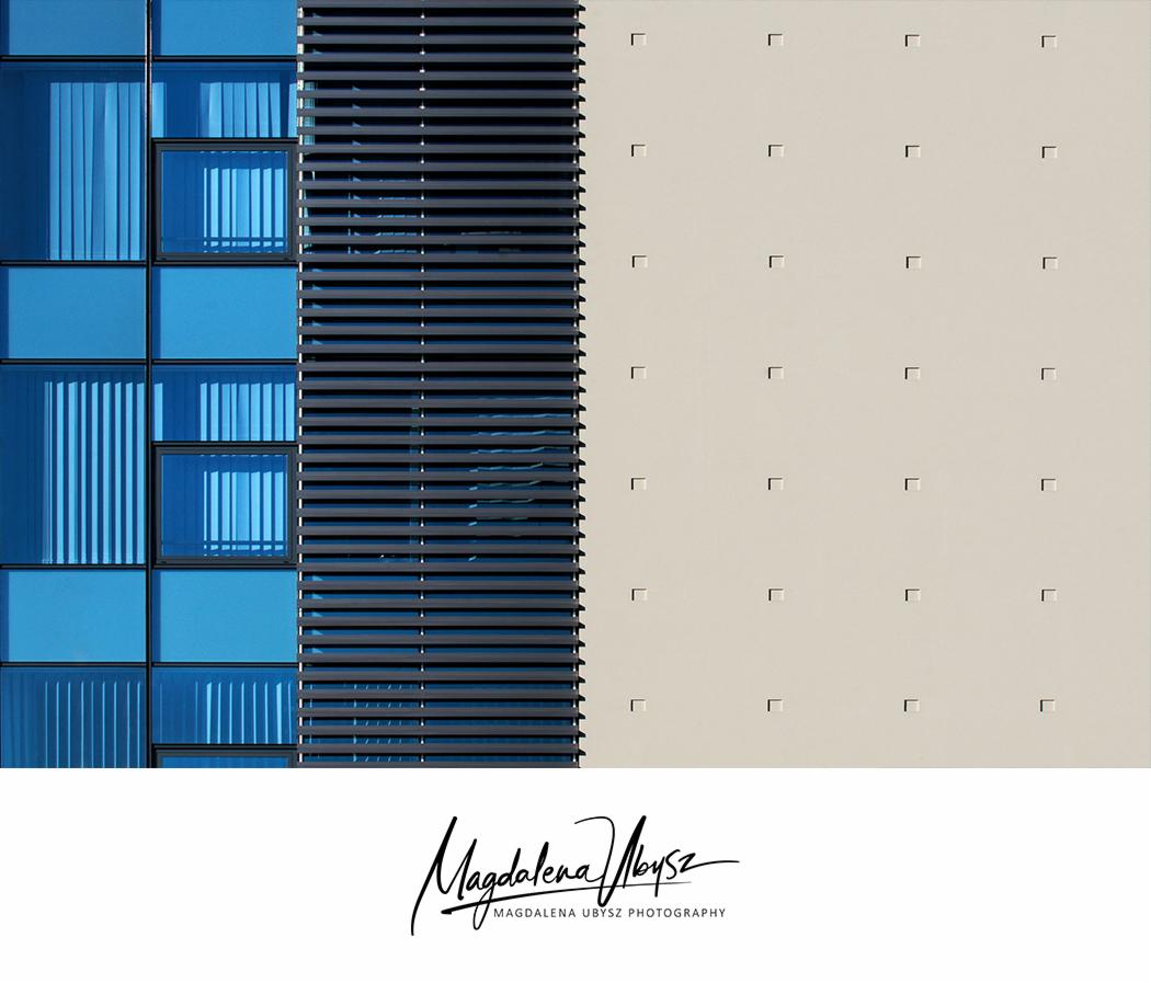 Fasada budynku z niebieskimi szklanymi panelami, żaluzjami i beżową ścianą z regularnymi wgłębieniami, sfotografowana z bliska.