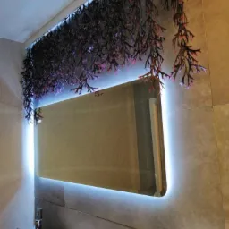 Nowoczesna łazienka z podświetlanym lustrem i dekoracyjnymi sztucznymi roślinami zwisającymi z sufitu, umywalka w kształcie misy i szara płytka na ścianach.