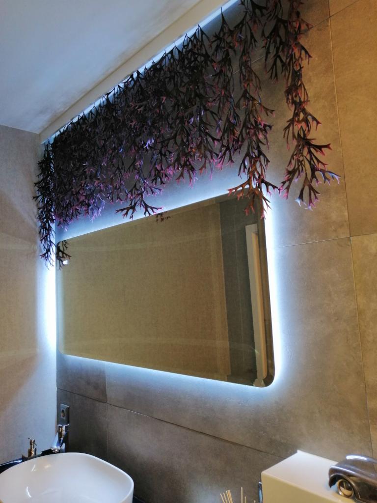 Nowoczesna łazienka z podświetlanym lustrem i dekoracyjnymi sztucznymi roślinami zwisającymi z sufitu, umywalka w kształcie misy i szara płytka na ścianach.
