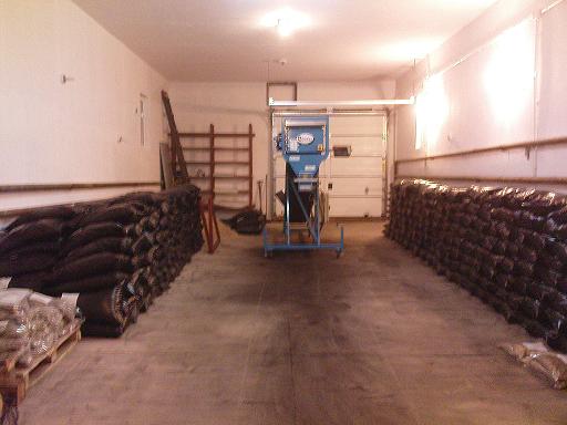 Magazyn z ułożonymi workami opału, w tle widoczna maszyna do pakowania i brama garażowa.