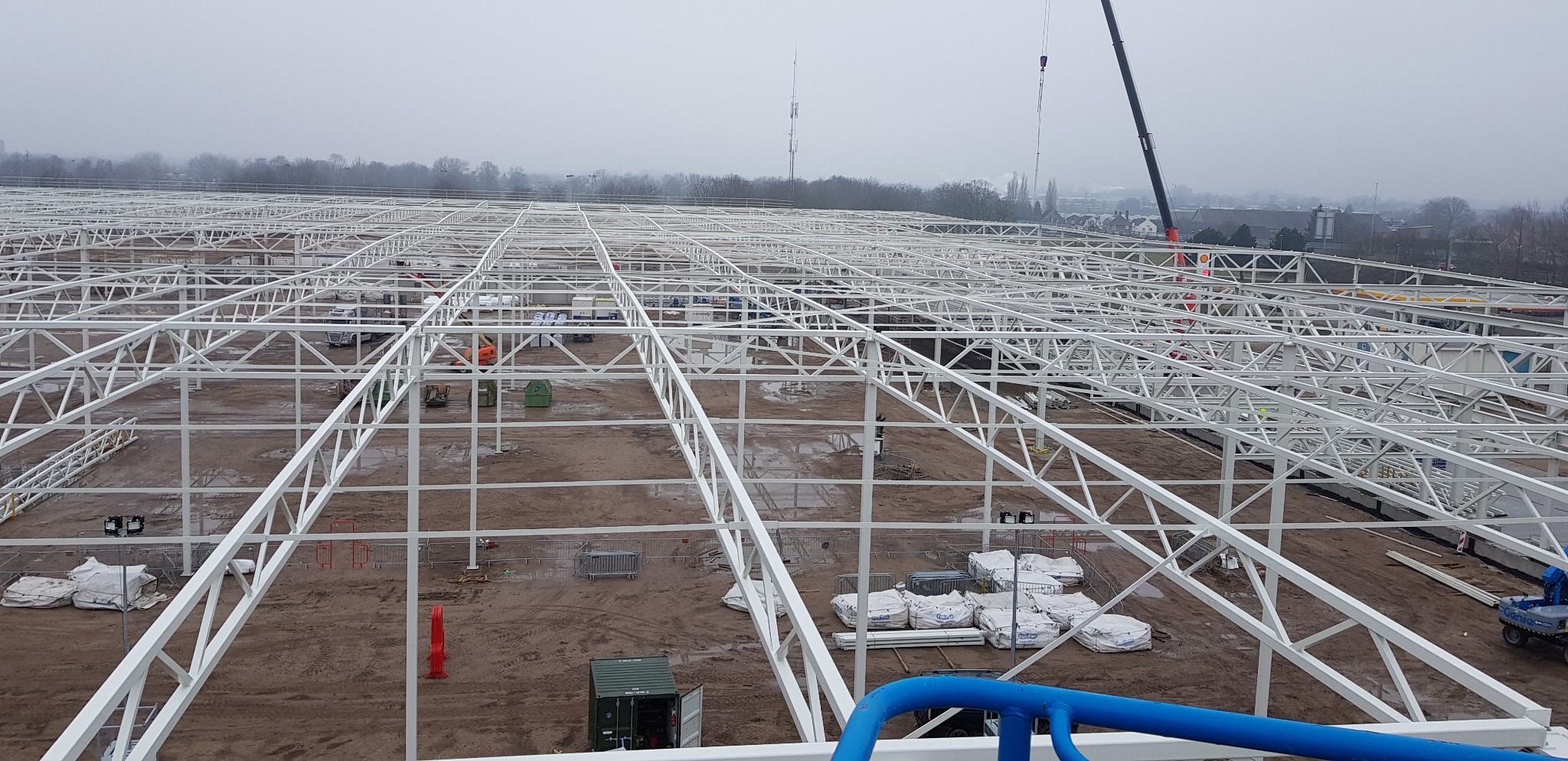 Rozległa, biała konstrukcja stalowa hali w trakcie budowy, widok z góry, z widocznymi elementami montażowymi, workami z materiałami budowlanymi i dźwigiem w tle, w pochmurny dzień.
