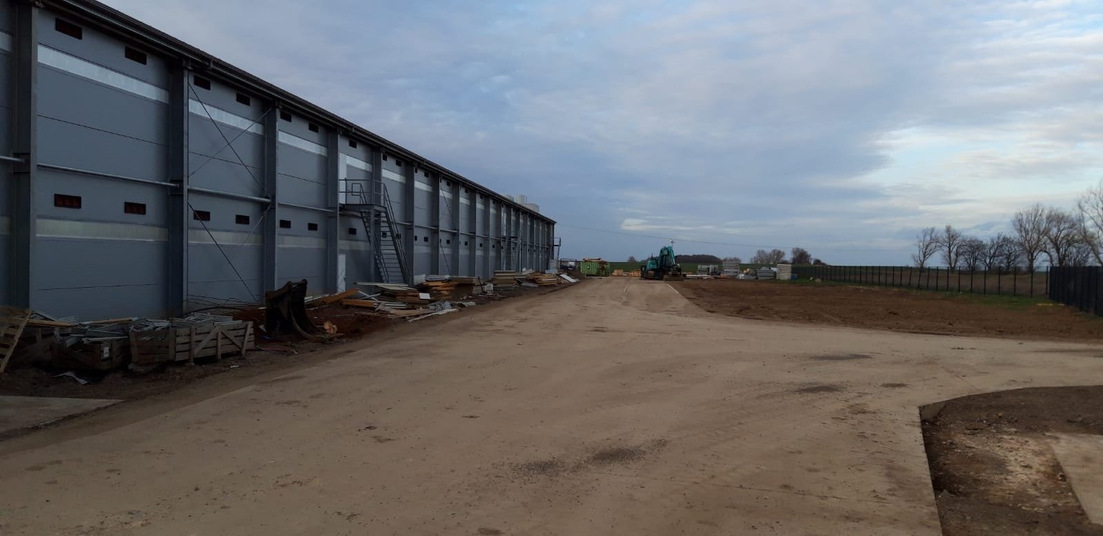 Budowa hali z szarej blachy trapezowej, widoczny teren budowy, sprzęt budowlany i składowane materiały.