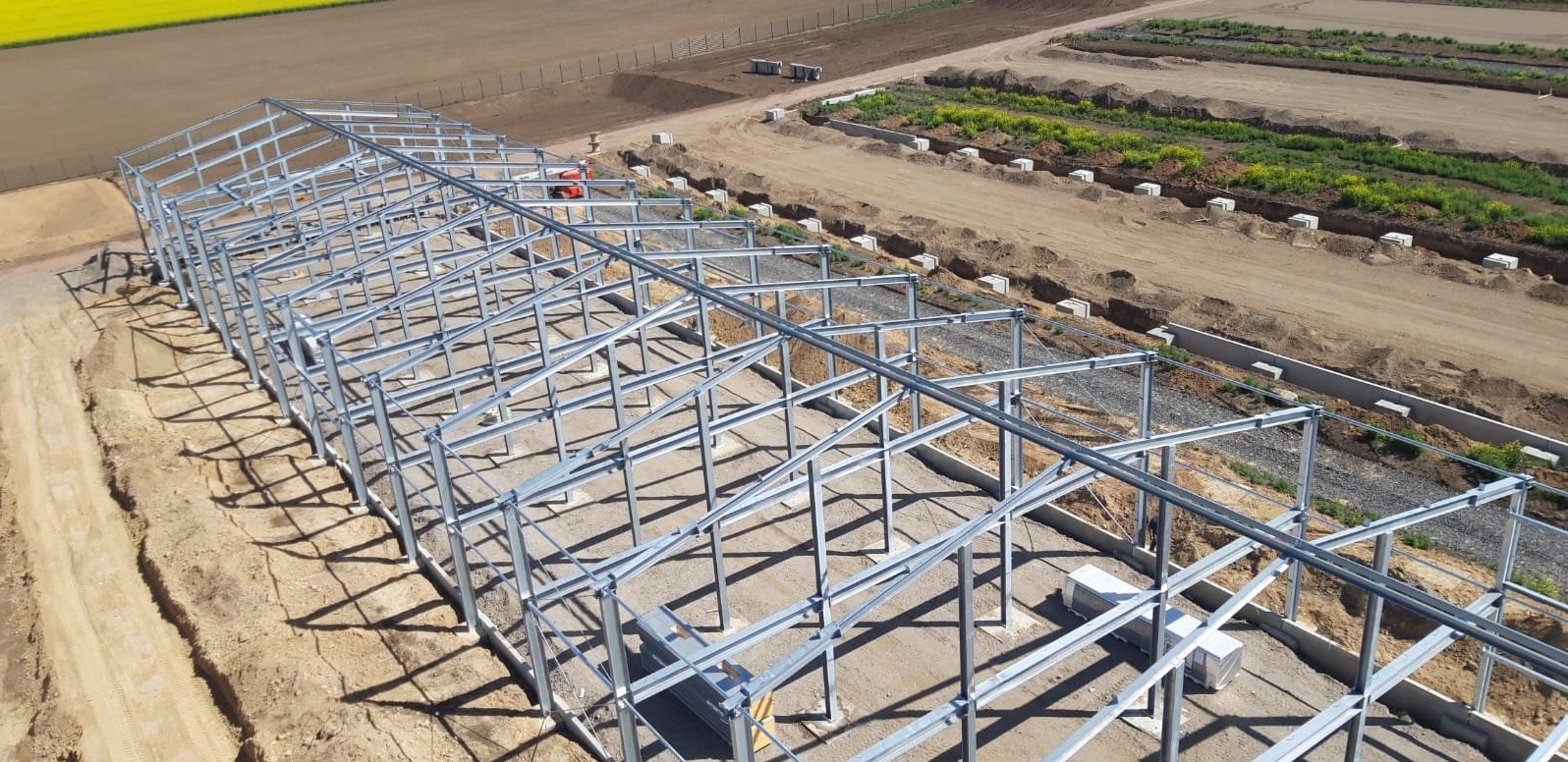 Widok z lotu ptaka na szkielet stalowej hali w trakcie budowy, widoczne fundamenty i przygotowane podłoże, w tle pola uprawne.