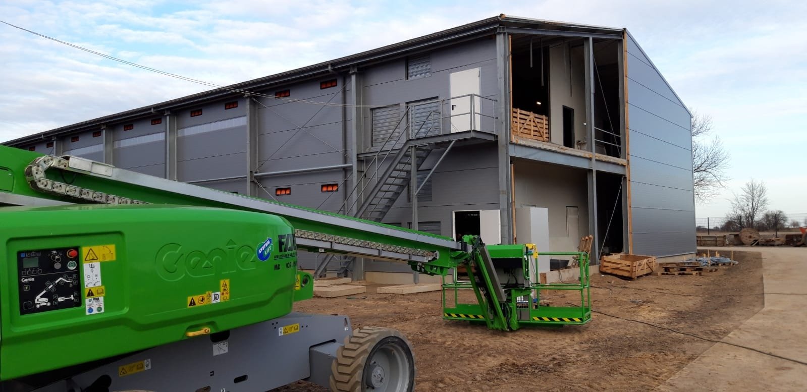 Montaż dwukondygnacyjnego budynku z paneli warstwowych, widoczny podnośnik koszowy Genie i stalowa konstrukcja szkieletowa w trakcie prac wykończeniowych.