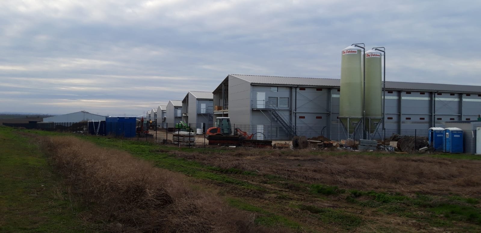 Wielkopowierzchniowy budynek gospodarczy z metalową konstrukcją, widoczne silosy, maszyny budowlane i przenośne toalety na tle pochmurnego nieba.