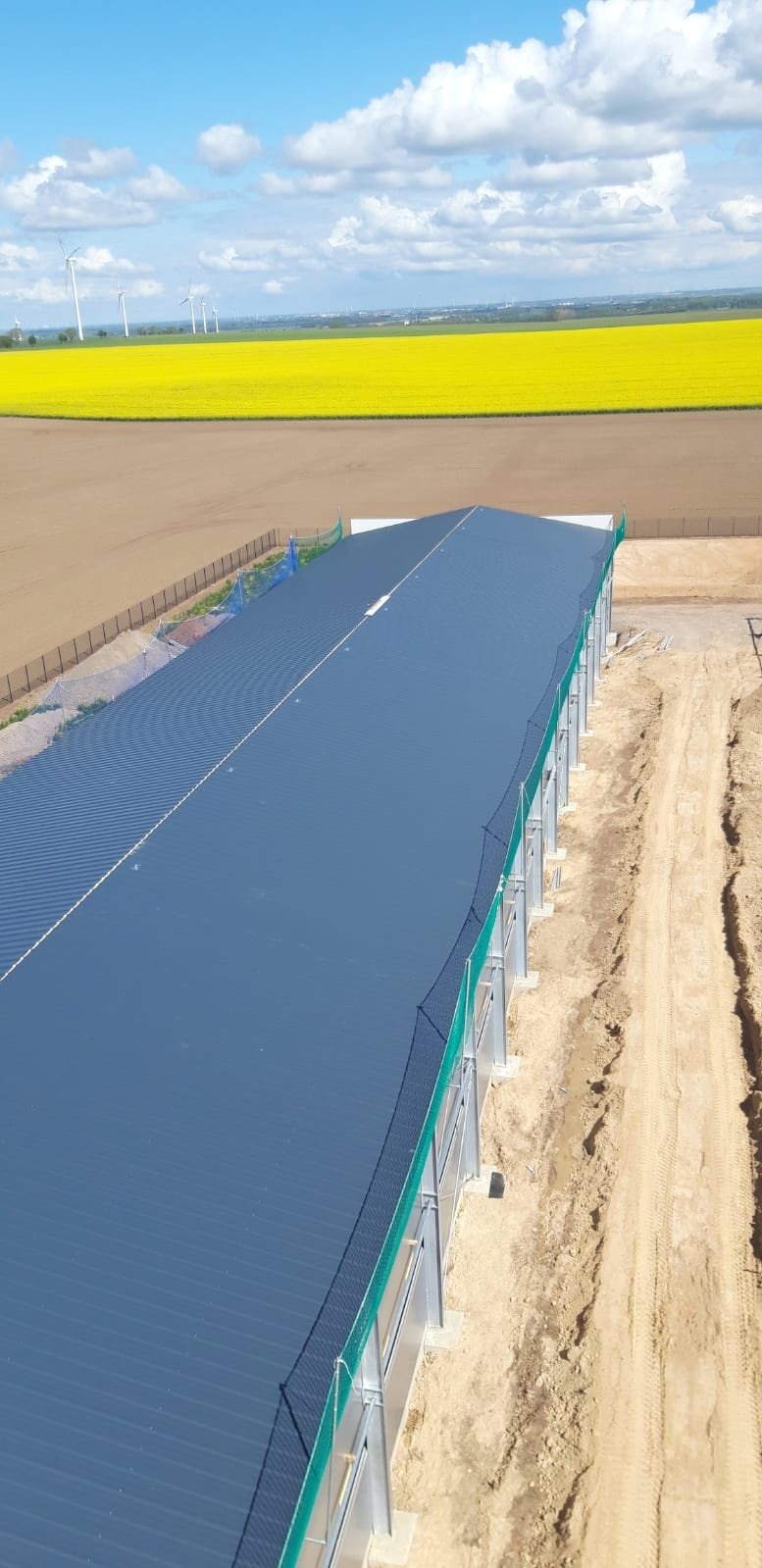 Szeroki kąt widzenia na budynek o stalowej konstrukcji z szarym dachem trapezowym, zabezpieczony siatką ochronną, na tle żółtego pola rzepaku i turbin wiatrowych.