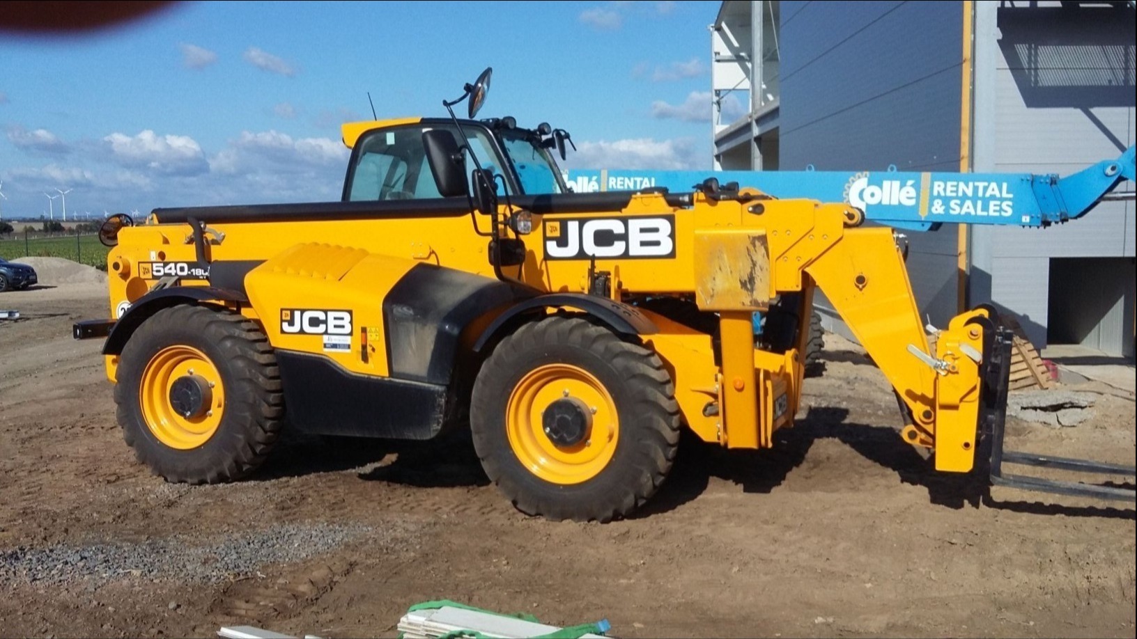 Żółta ładowarka teleskopowa JCB 540-180 w terenie budowy, widoczne w tle wiatraki i budynek w trakcie budowy.