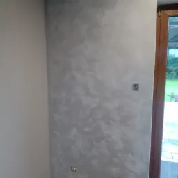 Ściana w rogu pomieszczenia pokryta dekoracyjną, szarą powłoką z efektem chmur, widoczne gniazdko elektryczne bez obudowy i fragment drewnianej ramy okiennej.