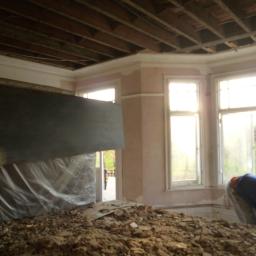 Łukasz Nawrocki - Demontaż starego parkietu w jasnym pokoju z oknami wykuszowymi, widoczne odsłonięte belki stropowe i fragment zabezpieczonej folią ściany.