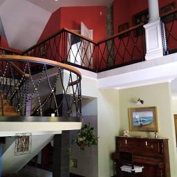 Łukasz Nawrocki - Dwupoziomowe wnętrze domu z drewnianymi schodami i dekoracyjną balustradą, czerwona ściana w tle, widoczny fragment osoby w niebieskiej koszulce.