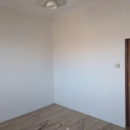 Pokój w trakcie układania paneli podłogowych, widoczne deski paneli, młotek i listwy przypodłogowe przy ścianie. Ściany pomalowane na jasny kolor, prosta lampa sufitowa.