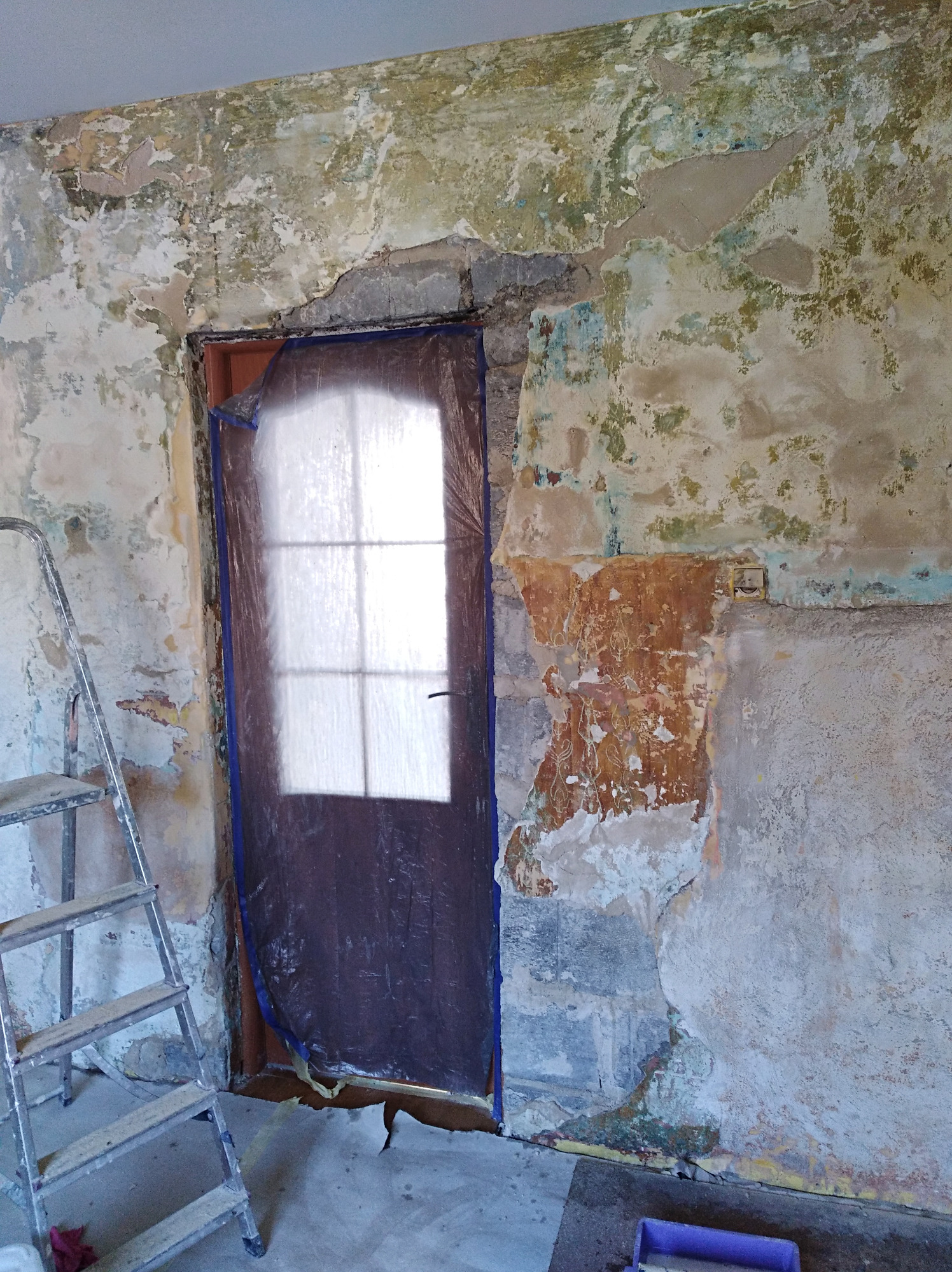 Przygotowanie ściany do prac wykończeniowych: zabezpieczone drzwi folią, odsłonięte warstwy starej farby i tynku, aluminiowa drabina w rogu.