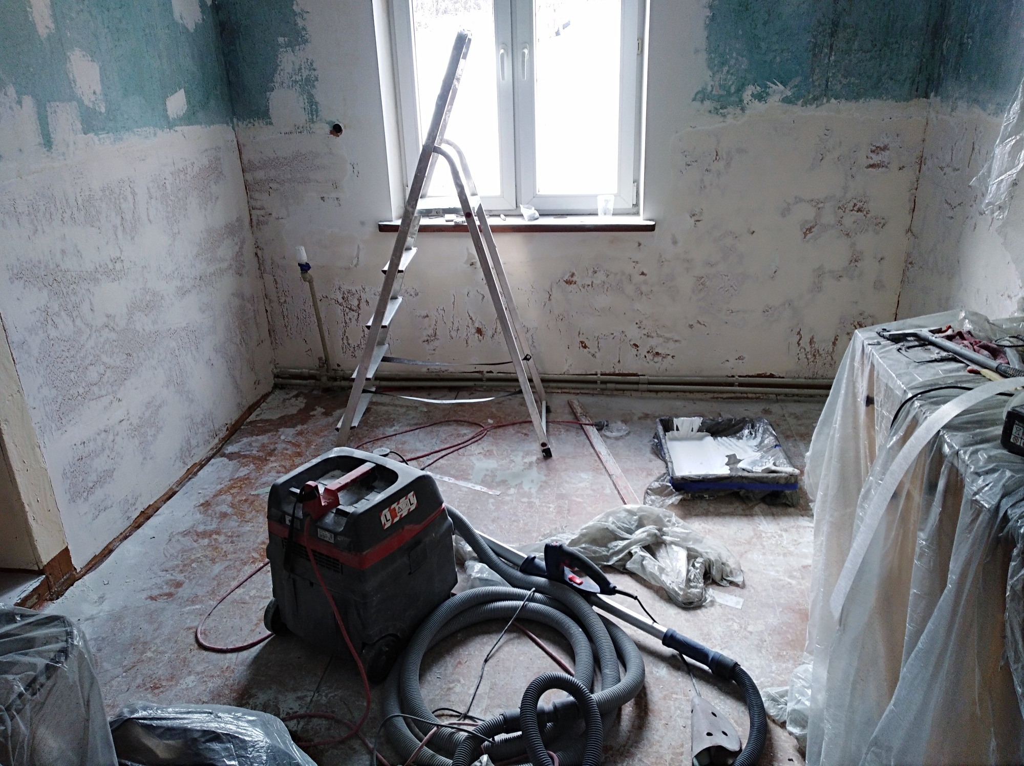Pokój w trakcie remontu, ściany częściowo przygotowane, widoczny odkurzacz przemysłowy, drabina aluminiowa i zabezpieczone folią meble.
