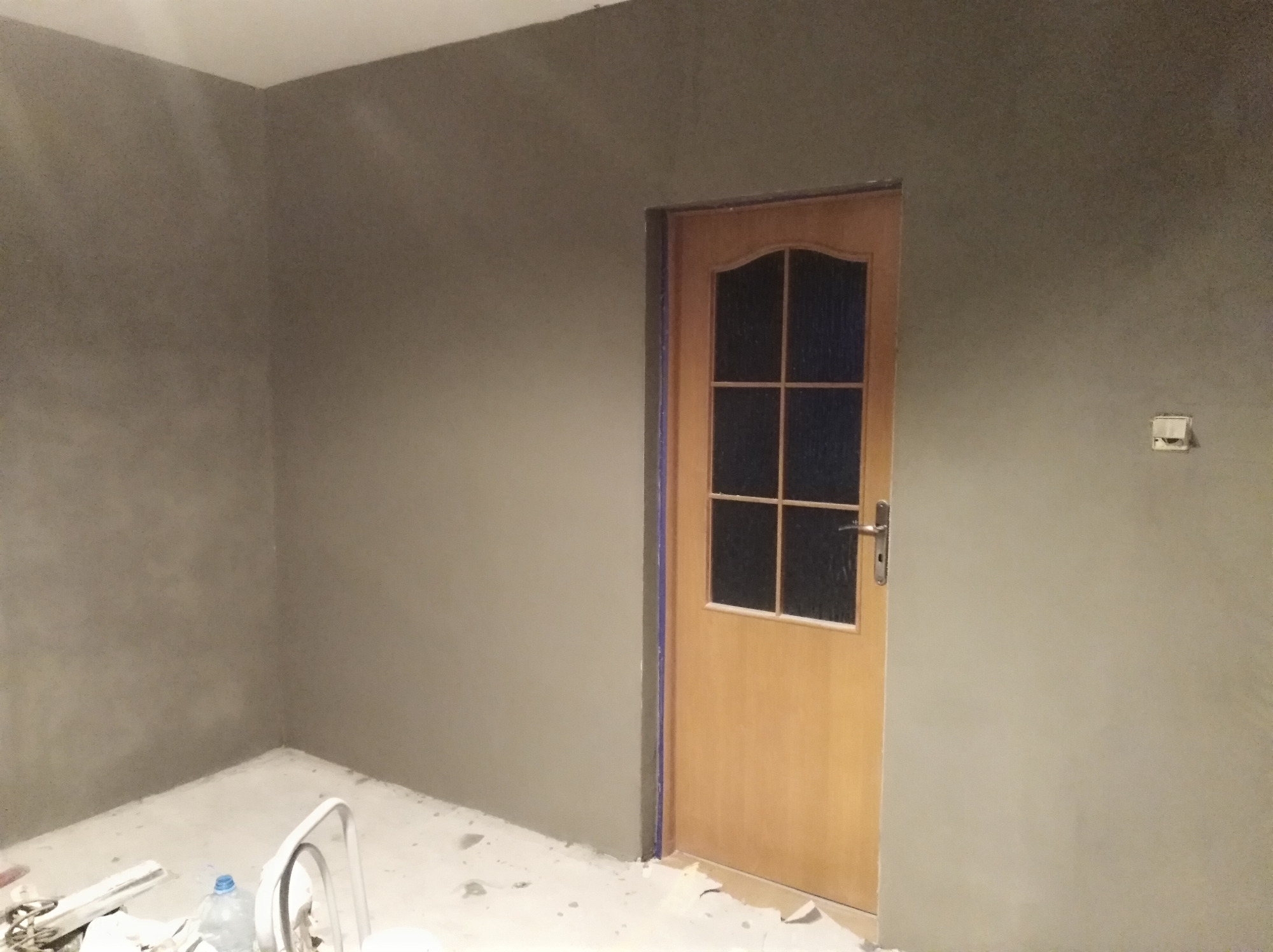 Pokój po nałożeniu szarej warstwy wykończeniowej na ścianach, widoczna drewniana ościeżnica drzwiowa z szybami i białe akcesoria malarskie na podłodze.