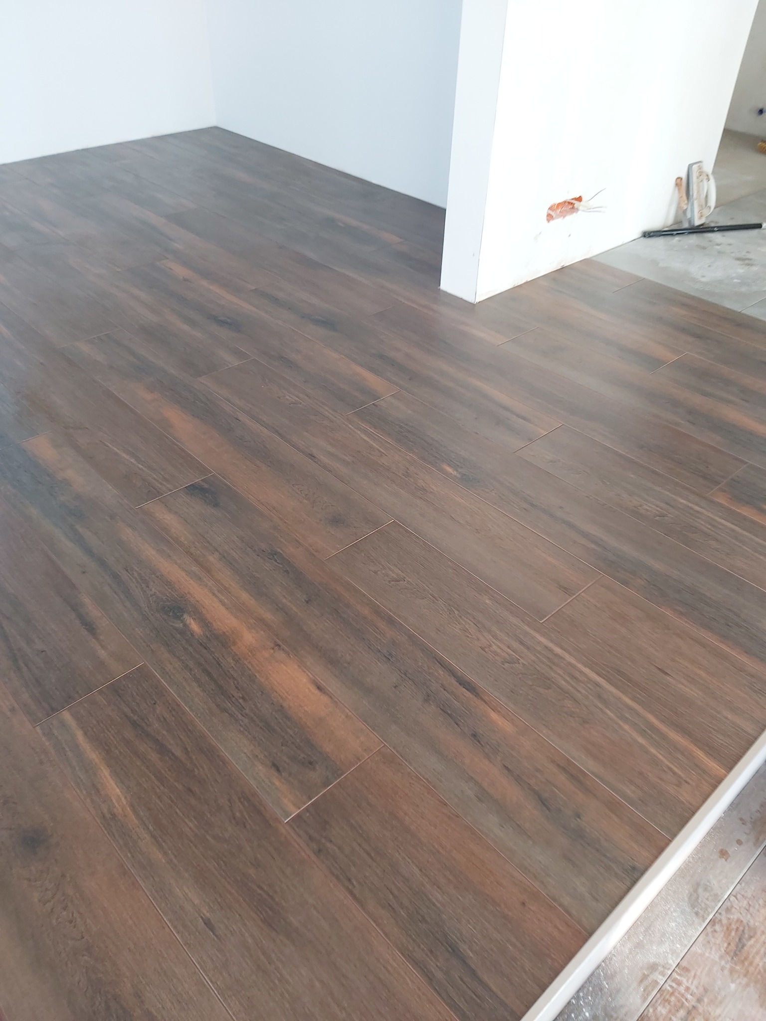 Ułożona podłoga z płytek imitujących drewno w odcieniach brązu i beżu, widoczne narzędzia i ślady prac wykończeniowych przy ścianie.