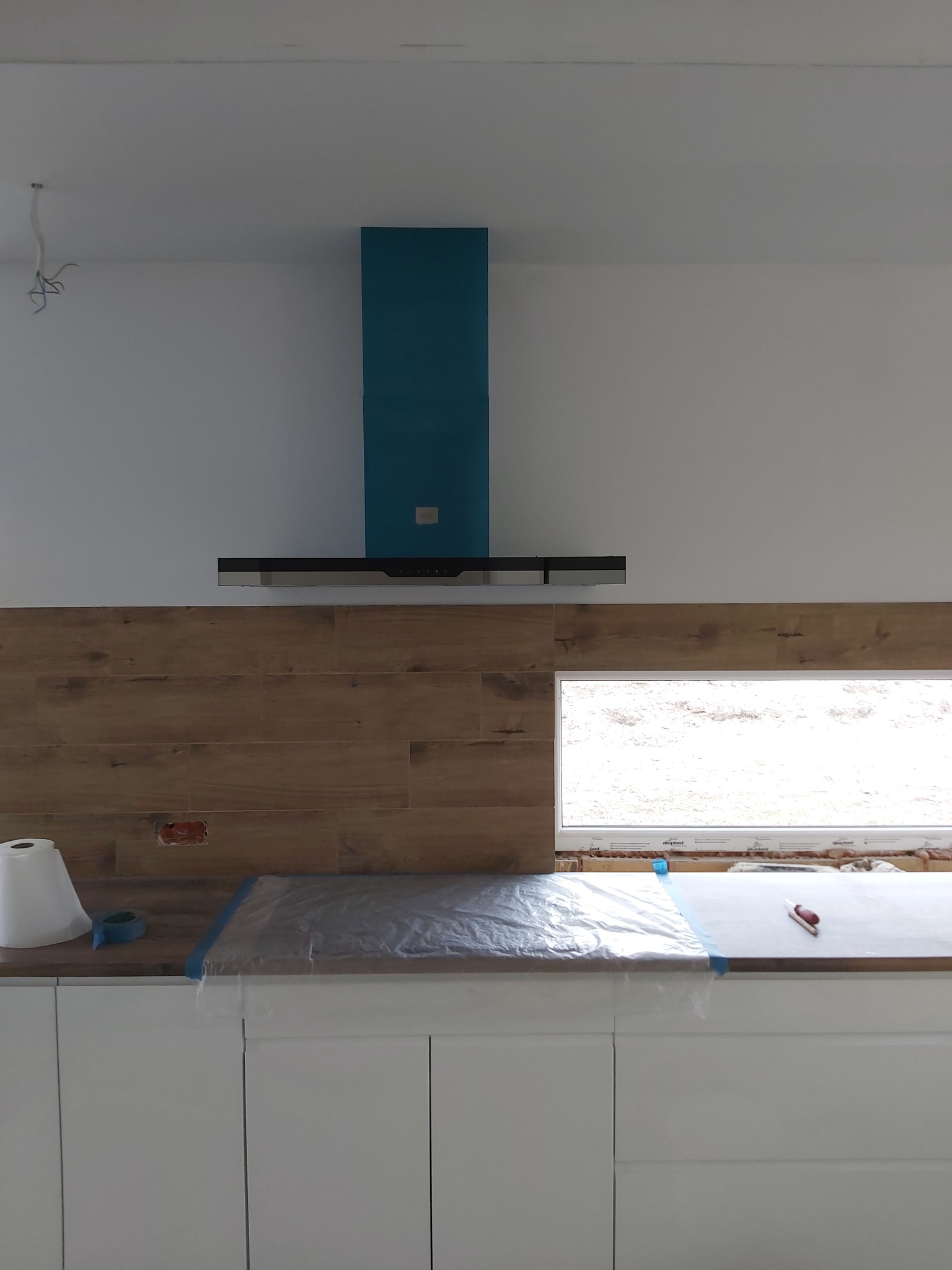 Montaż okładziny drewnopodobnej na ścianie w kuchni, widoczny okap w kolorze turkusowym, zabezpieczony blat i odkryta puszka elektryczna.