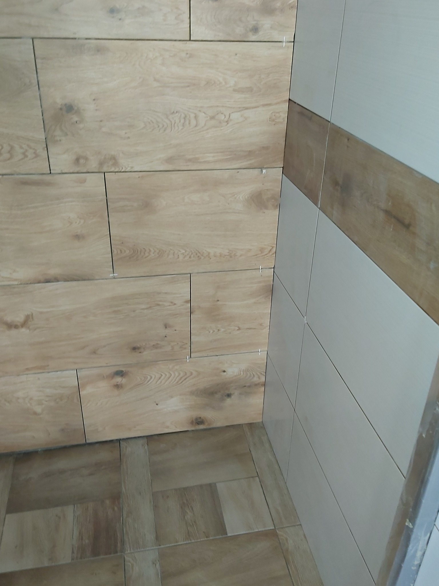 Ściana i podłoga wyłożone płytkami imitującymi drewno, z krzyżykami dystansowymi w fugach, kontrastujące z białymi płytkami z brązowym pasem na sąsiedniej ścianie.