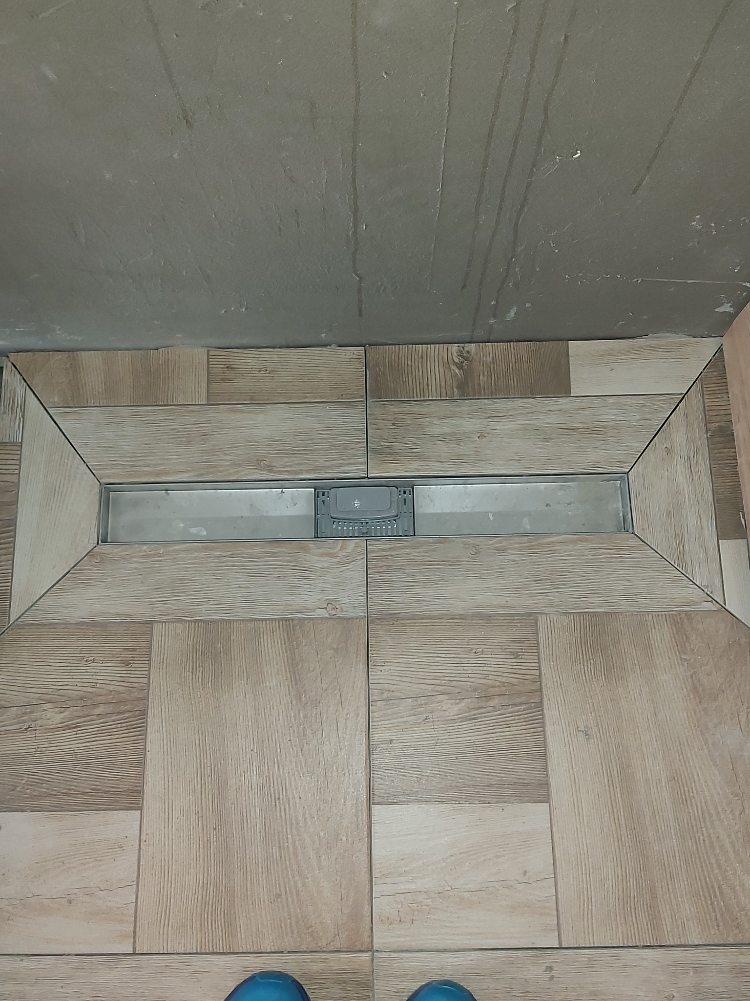 Ułożona podłoga z płytek imitujących drewno wokół liniowego odpływu prysznicowego; widoczne ślady po kleju na ścianie w tle.