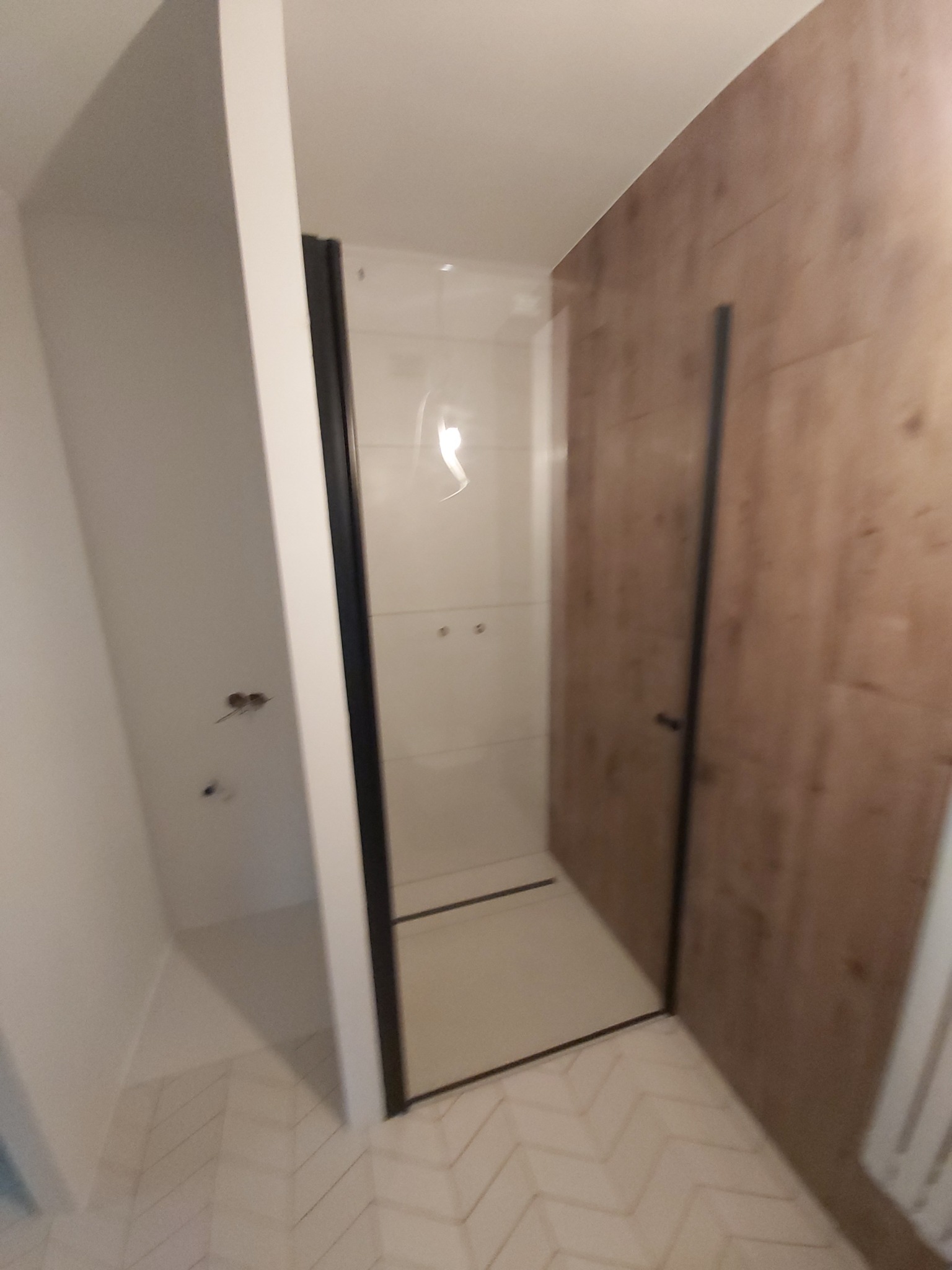 Nowoczesna kabina prysznicowa z czarnymi profilami, jasnymi płytkami wewnątrz i okładziną imitującą drewno na ścianie, widoczna instalacja elektryczna obok kabiny.