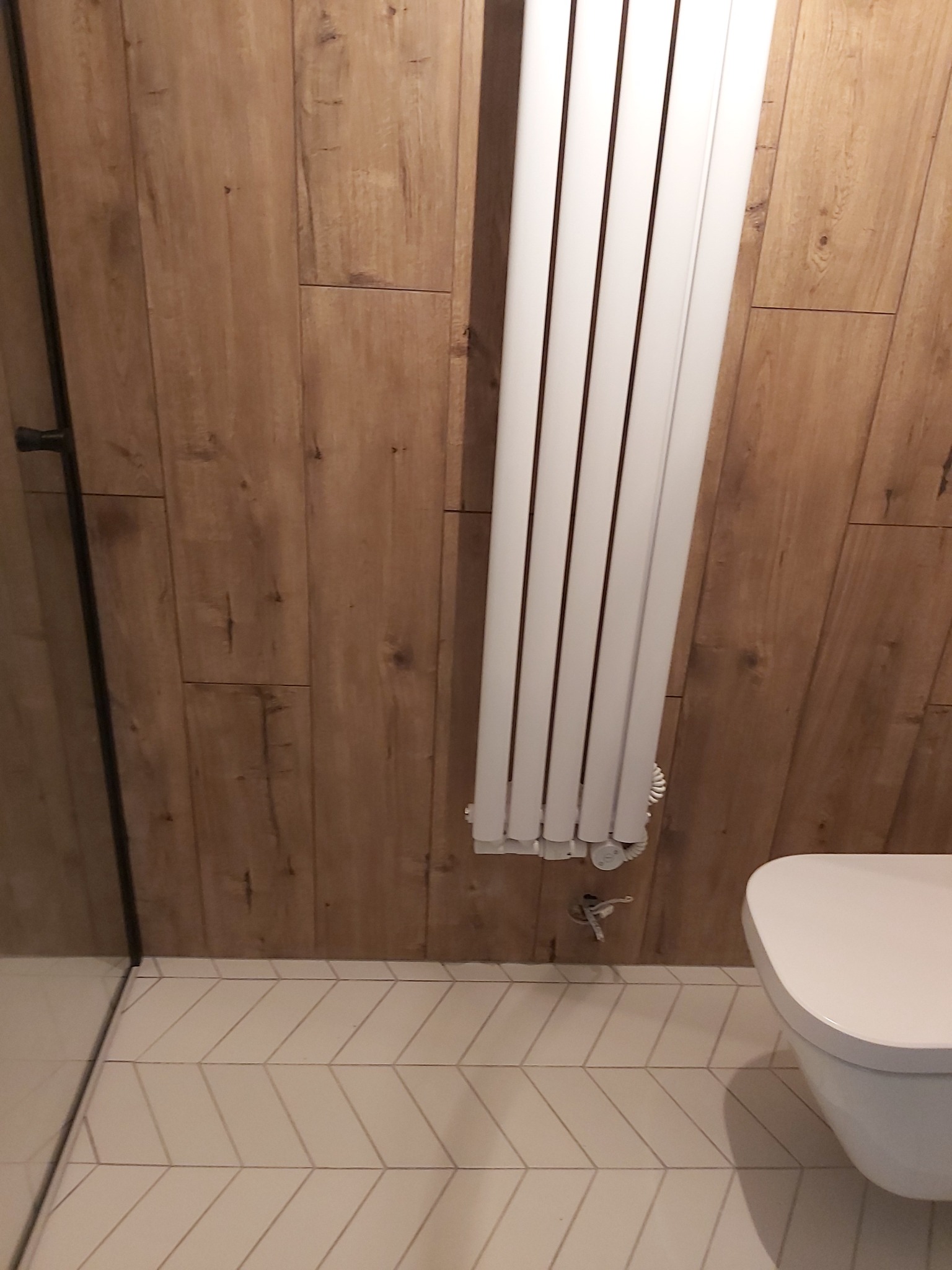 Nowoczesna łazienka z drewnopodobnymi płytkami ściennymi, białym grzejnikiem drabinkowym i toaletą wiszącą na tle kremowych płytek podłogowych ułożonych we wzór jodełki.