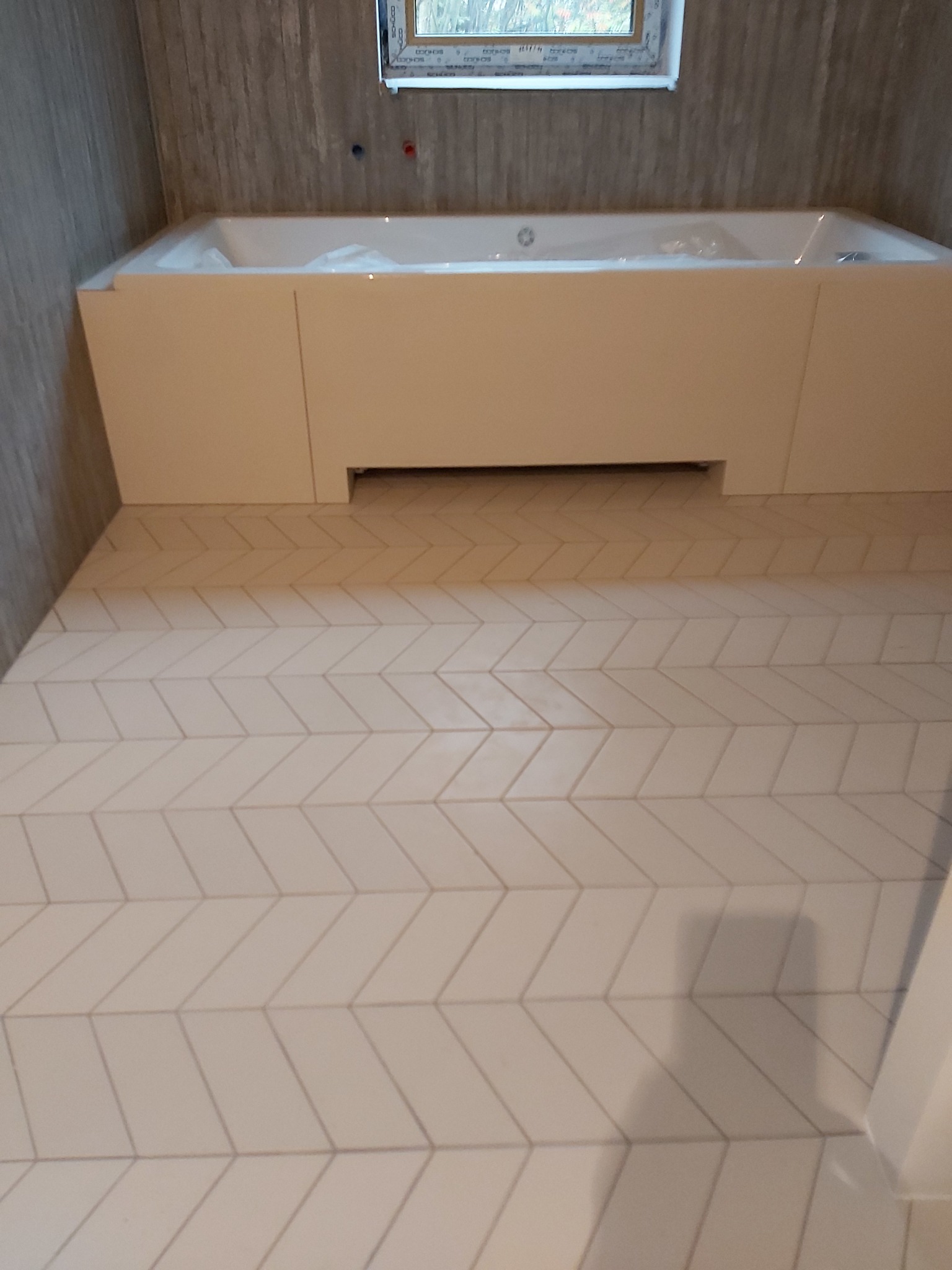 Biała wanna obudowana płytami MDF i podłoga wyłożona jasnymi płytkami w jodełkę, widok na fragment łazienki z oknem.