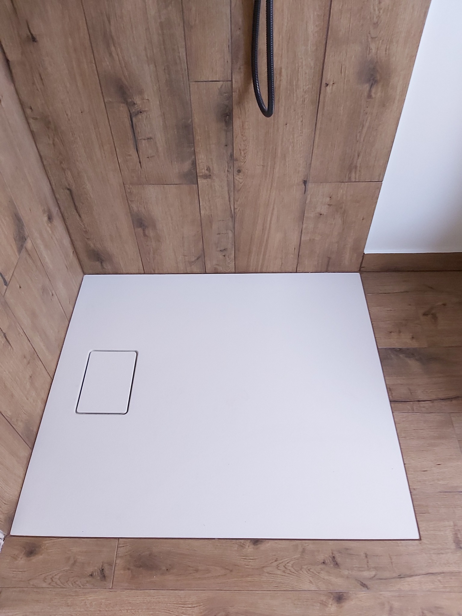 Biały, prostokątny brodzik z odpływem w rogu, otoczony drewnopodobnymi płytkami ściennymi i podłogowymi, z widocznym czarnym wężem prysznicowym.