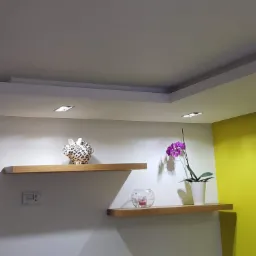 Pokój z podwieszanym sufitem z wbudowanym oświetleniem punktowym, drewnianymi półkami z dekoracjami i ścianą pomalowaną na biało i limonkowo.