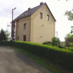 Dwupiętrowy dom z beżową elewacją i ciemnym dachem, widziany z dołu, z widocznymi rusztowaniami z boku i schludnym żywopłotem na pierwszym planie.