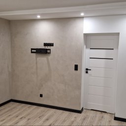 Marcin Szopa PROREMDOM - Ściana z dekoracyjnym tynkiem w odcieniach beżu, z zamontowanym uchwytem pod telewizor, białe drzwi z poziomymi czarnymi paskami, widoczne fragmenty surowej ściany po lewej stronie drzwi.