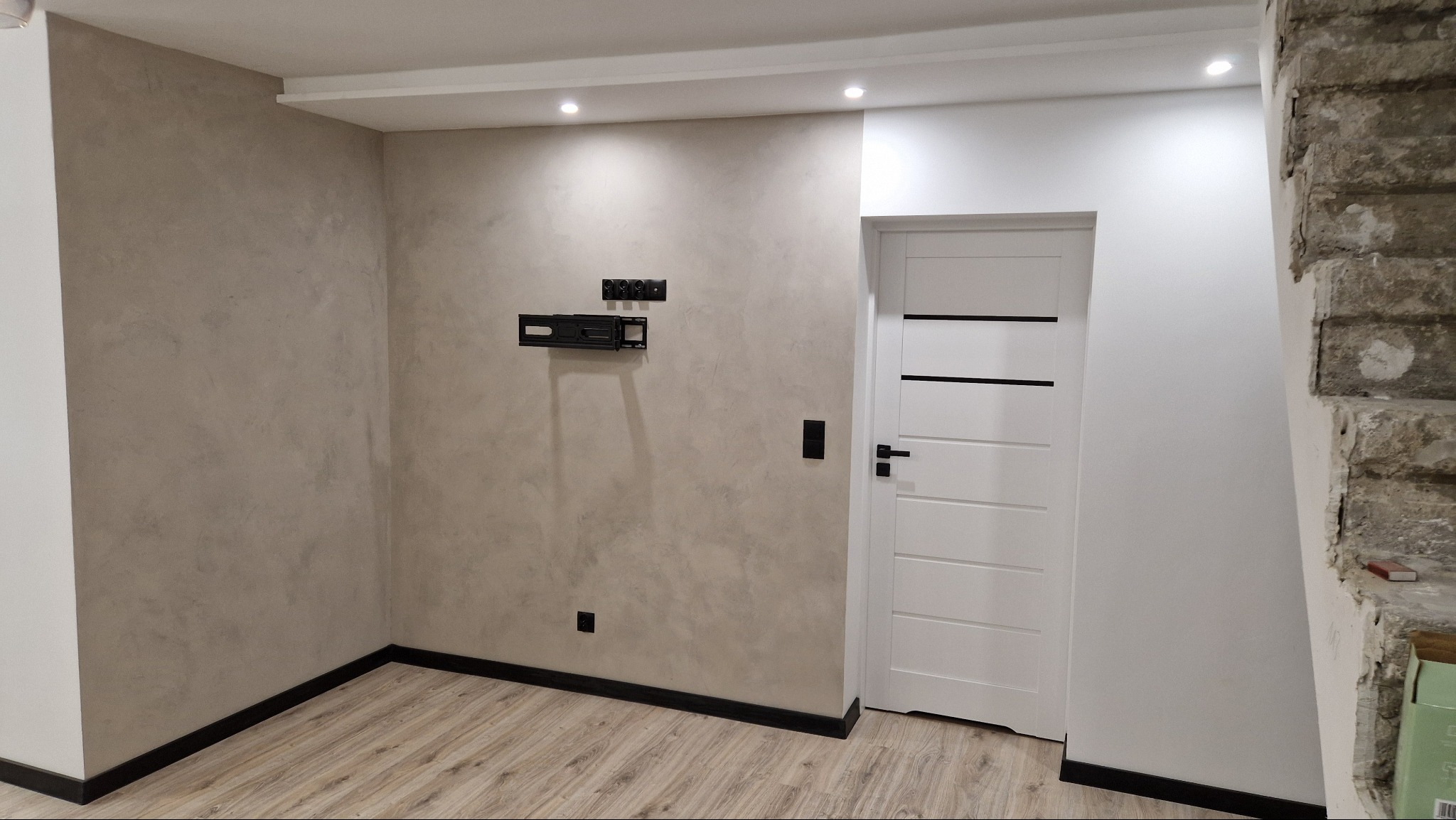 Ściana z dekoracyjnym tynkiem w odcieniach beżu, z zamontowanym uchwytem pod telewizor, białe drzwi z poziomymi czarnymi paskami, widoczne fragmenty surowej ściany po lewej stronie drzwi.